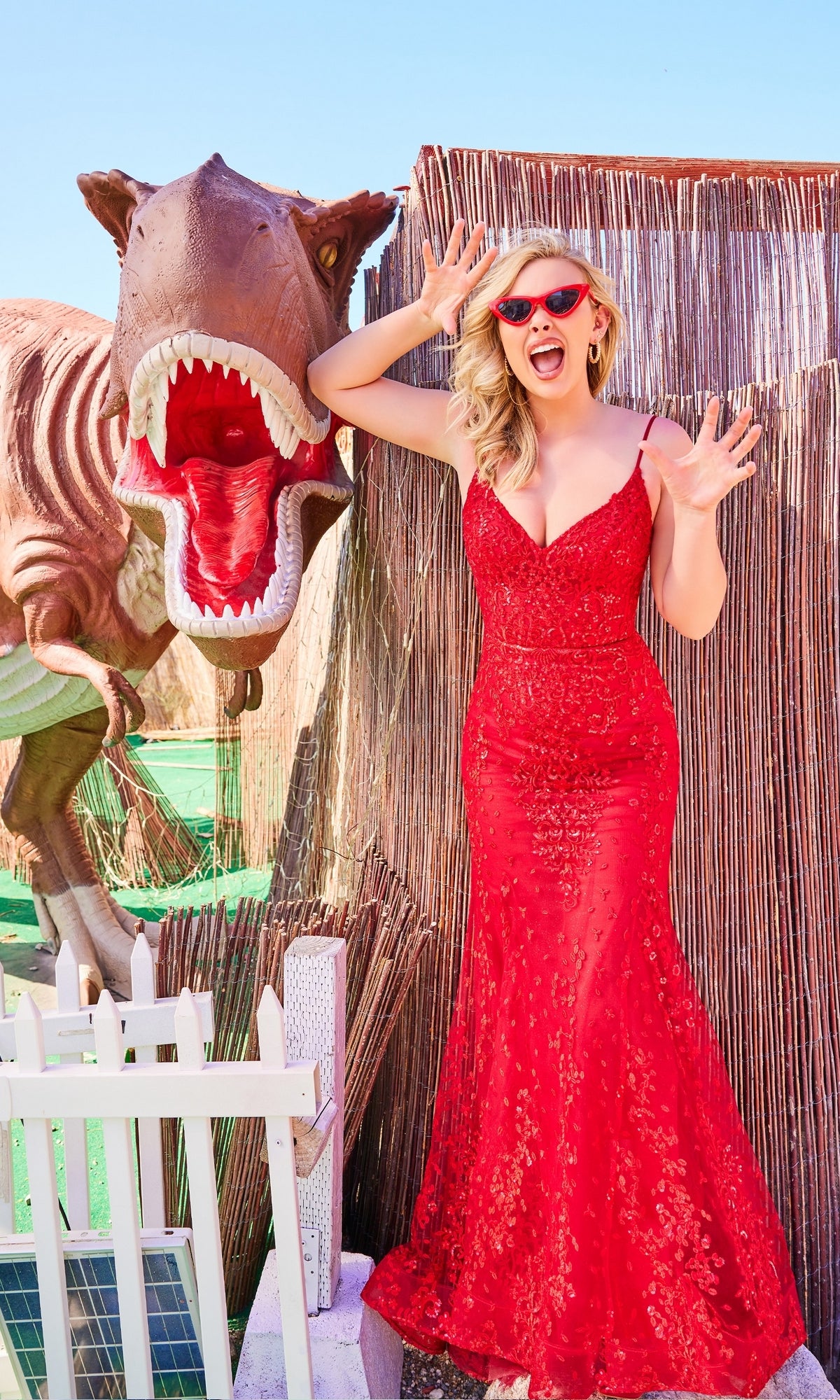 A woman in a Sequin-Lace Open-Back Long Mermaid Prom Dress and red sunglasses poses with her hands raised beside a large dinosaur model with its mouth open.